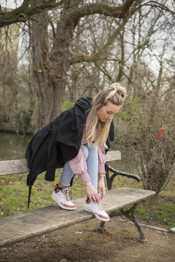 Eine Frau sitzt auf einer Holzbank in einem Park und richtet ihre Barfußschuh-Schnürsenkel. Sie trägt einen schwarzen Mantel, den pinkfarbenen Pullover Rosa Waterproof aus der VAQATION-Linie von ZAQQ, hellblaue Jeans und weiße Sneakers. Den Hintergrund bilden spärliche Bäume und Büsche.