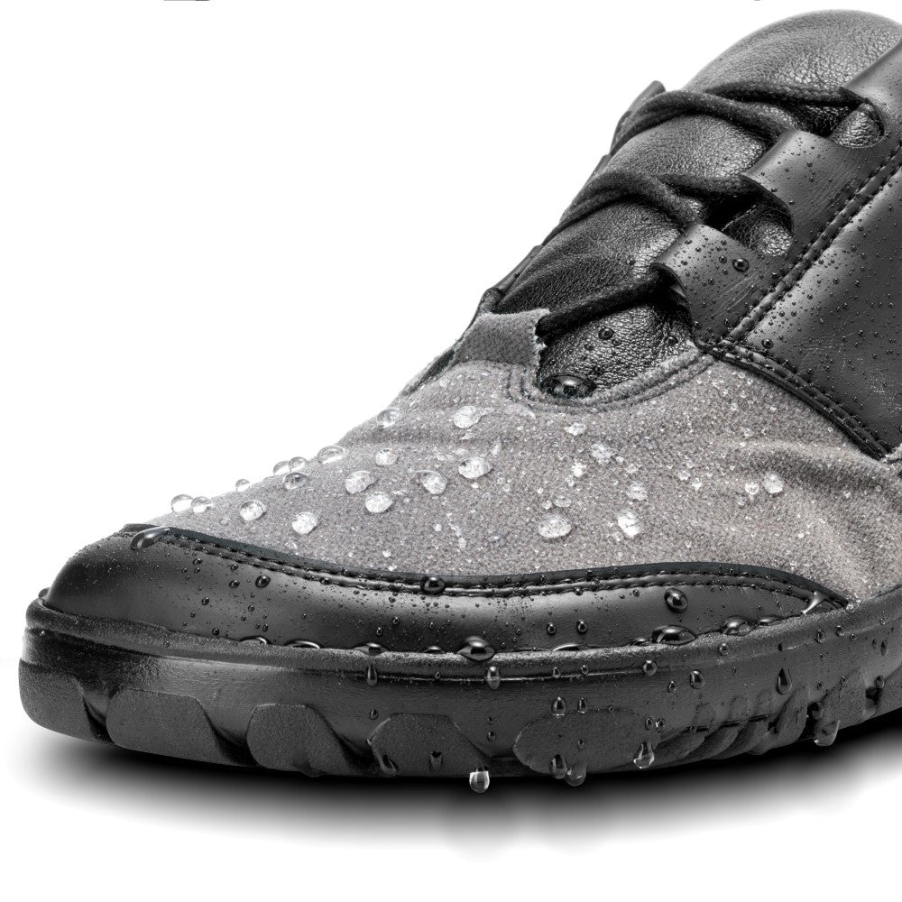 Nahaufnahme eines QUEST Low Black Waterproof-Schuhs von ZAQQ mit Wassertropfen auf seiner Oberfläche. Der Schuh hat ein strukturiertes graues Obermaterial mit schwarzen Lederakzenten und eine robuste schwarze Sohle. Die Wassertropfen betonen sein wasserdichtes Design und machen ihn ideal für Abenteuer im Freien.